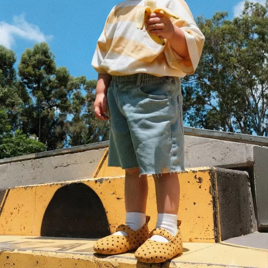 A child standing outdoors on yellow concrete steps under a blue sky, wearing a cream and yellow tie-dye long-sleeve top, light blue denim shorts, white socks, and yellow polka-dot shoes.